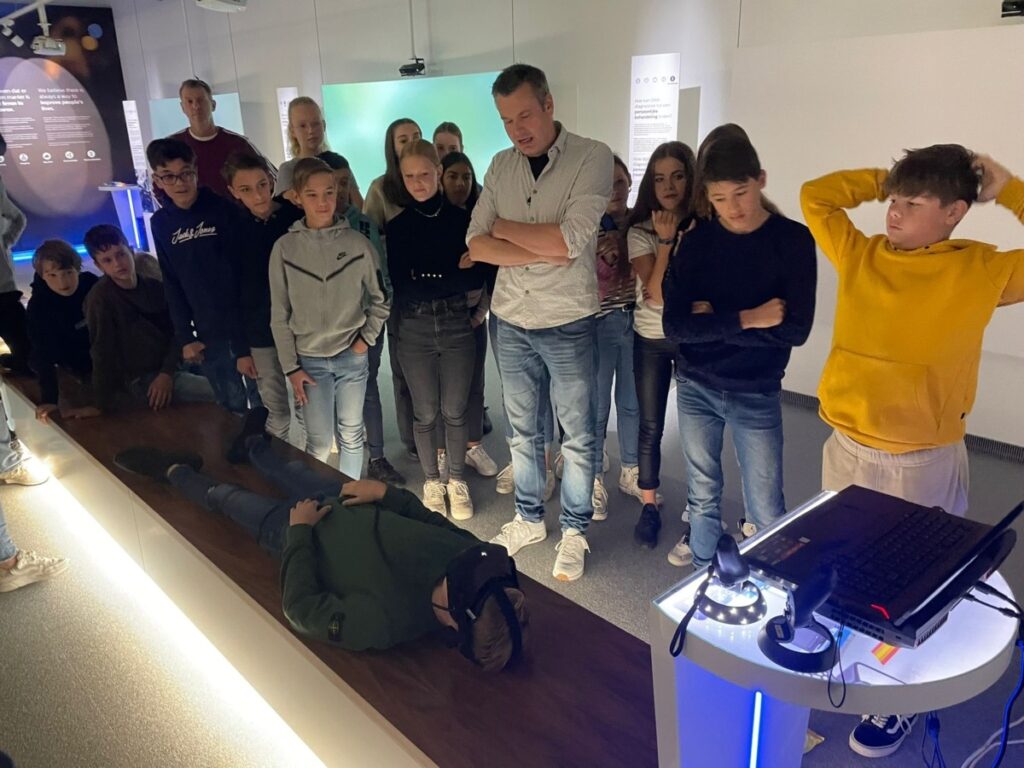 Leerlingen kijken naar een leerling met een VR-bril op die op de grond ligt.