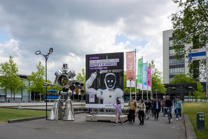 Bezoekers lopen buiten op de campus van Universiteit Twente langs reclame voor het nieuwe Robotics Centre